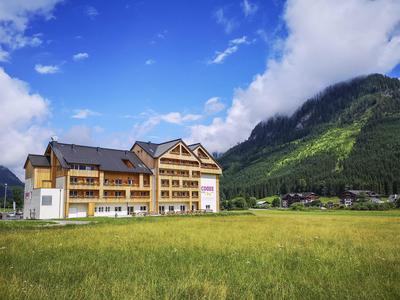 Großes gelbes Gebäude mit Holzdetails vor grüner Berglandschaft und blauem Himmel mit Wolken.