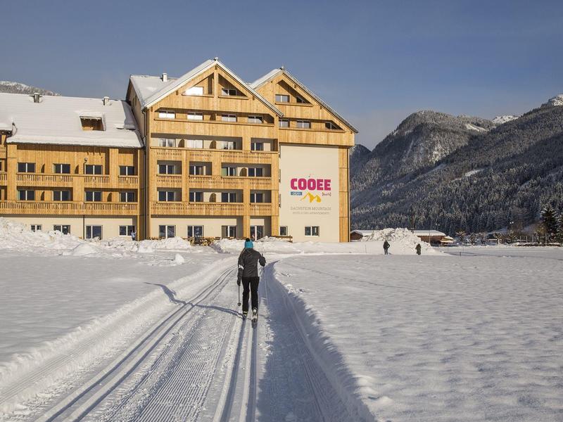 Skifahrer auf Loipe vor großem Hotel im verschneiten Bergtal bei klarem Himmel