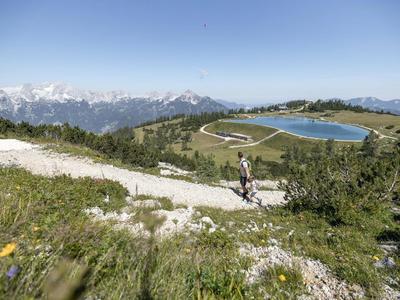 Berglandschaft mit Wanderweg, grünen Wiesen, Solarseen und schneebedeckten Gipfeln im Hintergrund.