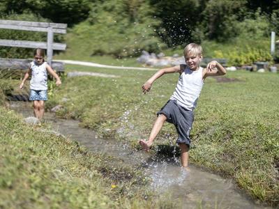 Zwei Kinder spielen lachend und springen barfuß in einem flachen Bach in einer grünen Wiese.