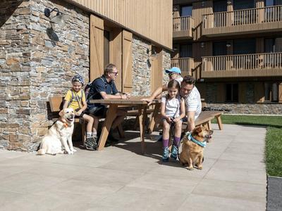 Famille avec deux enfants et deux chiens assis devant un hôtel de village alpin au soleil.