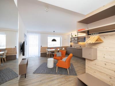 Moderner Wohnraum mit orangenen Sesseln, Holzwänden, Esstischen und grauem Teppich auf Holzboden.