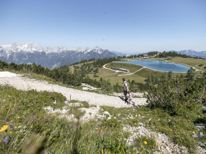Berglandschaft mit Wanderweg, grünen Wiesen, Solarseen und schneebedeckten Gipfeln im Hintergrund.