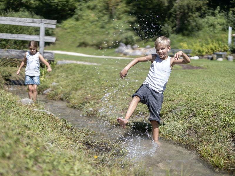 Zwei Kinder spielen lachend und springen barfuß in einem flachen Bach in einer grünen Wiese.