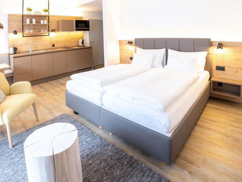 Helles Zimmer mit Doppelbett, Küchenzeile, zwei Sesseln, grauem Teppich und Holzboden.