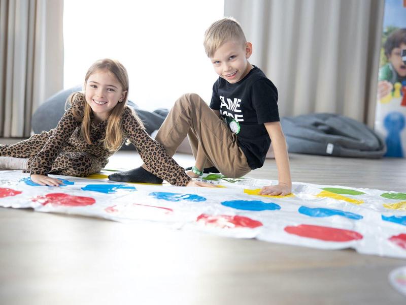 Zwei Kinder spielen auf einem bunten Twister-Spielteppich in einem hellen, modernen Raum.