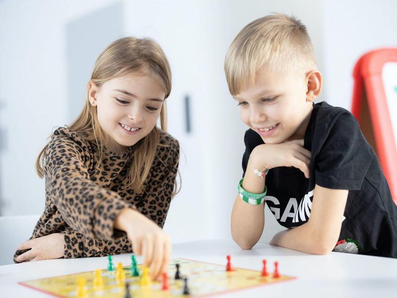 Zwei Kinder spielen fröhlich ein Gesellschaftsspiel mit bunten Spielfiguren am weißen Tisch.