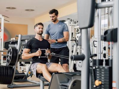 Zwei Männer trainieren im Fitnessstudio an einer Kraftmaschine, einer sitzt und zieht, der andere hilft.