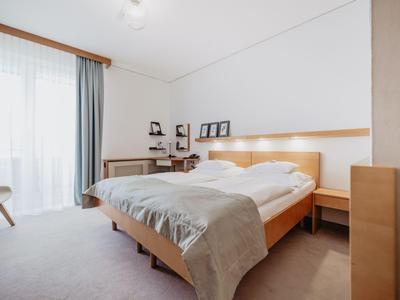 Helles, modernes Hotelzimmer mit Doppelbett, grauer Bettdecke und großem Fenster mit Vorhängen.