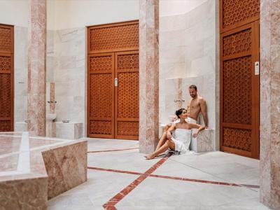 Luxuriöse Sauna mit hellen Marmorwänden, großen Holztüren und zwei entspannten Personen.