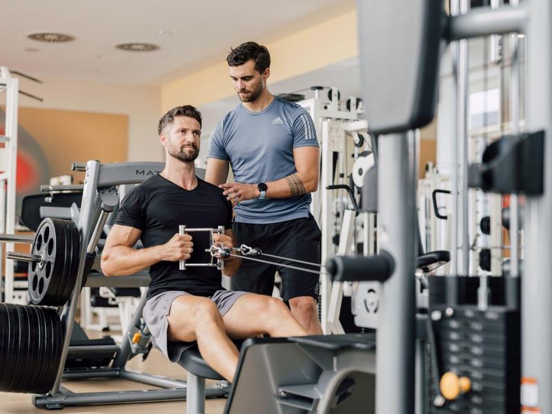 Zwei Männer im Fitnessstudio, einer trainiert Rudermaschine, der andere unterstützt ihn.