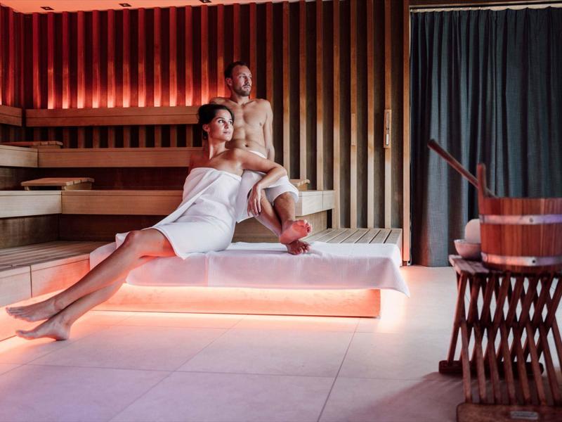 Zwei Frauen in weißen Handtüchern entspannen auf beleuchteten Holzliegen in moderner Sauna.