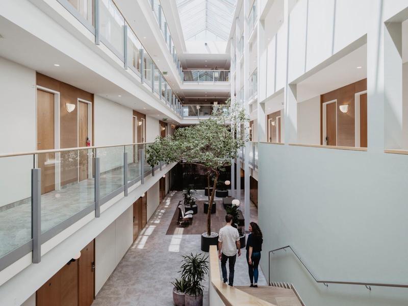 Hell gestaltetes Atrium mit Glasdach, zwei Etagen, Pflanzen und Personen im modernen Gebäude.