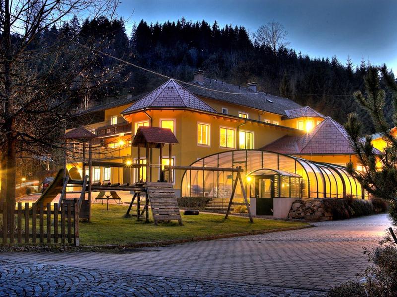 Großes beleuchtetes Haus mit Wintergarten und Umzäunung, umgeben von Bäumen bei Abenddämmerung.