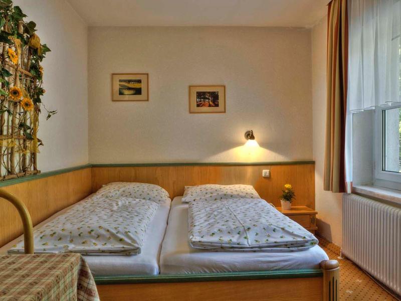 Kleines Einzelzimmer mit zwei Betten, Holzvertäfelung, Bildern an der Wand und einem Fenster mit Vorhang.