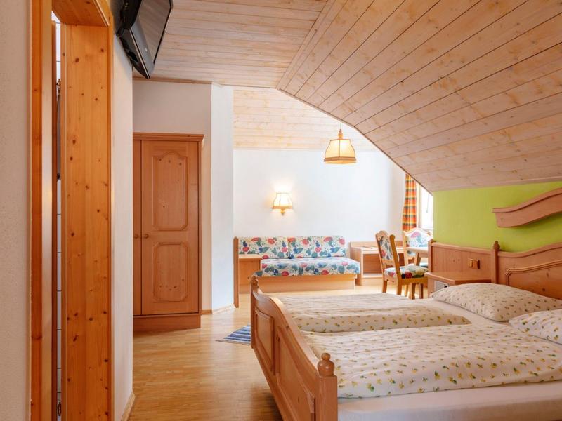 Gemütliches Zimmer mit Holzbett, schräger Holzdecke und Blick in einen Essbereich mit buntem Sofa.