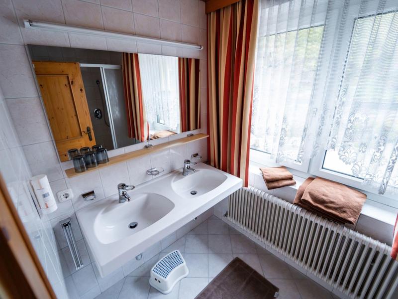 Helles Badezimmer mit Doppelwaschbecken, großem Spiegel, weißen Fliesen und Vorhängen am Fenster.