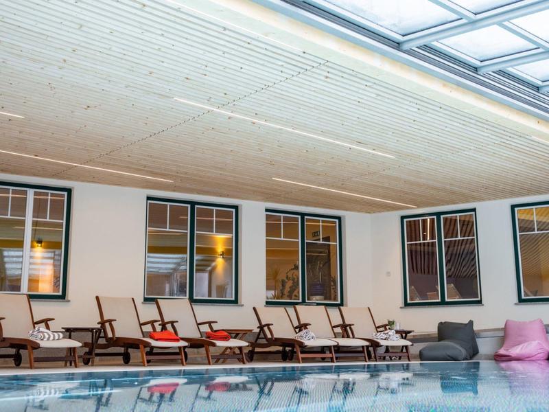 Innenpoolbereich mit Holzdecke, braunen Liegestühlen und rosa Sitzsäcken neben großem Fenster.