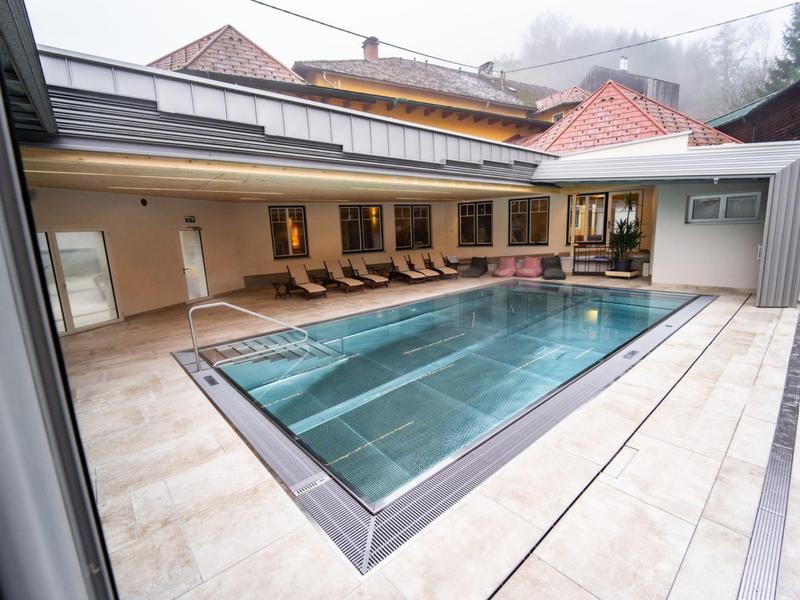 Moderner Innenpool mit Glaswand, Holzliegen und hellbeigen Fliesen in alpiner Umgebung.