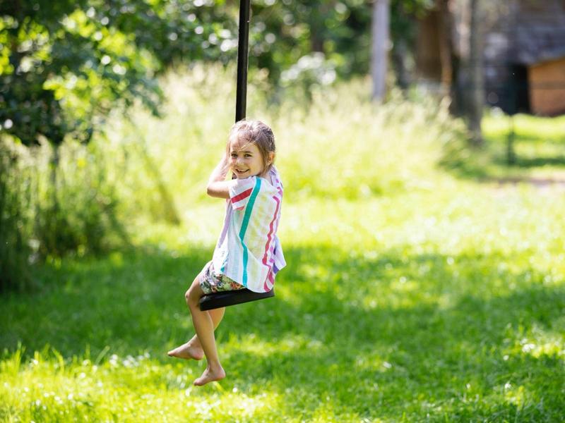 Mädchen schaukelt auf Seilschaukel im sonnigen Garten mit grünem Gras und Bäumen im Hintergrund.
