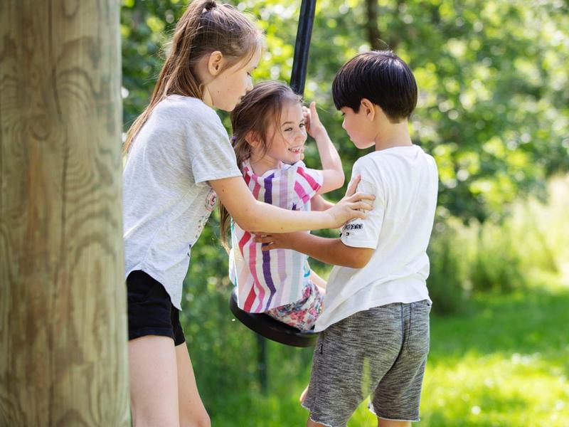 Drei Kinder spielen draußen auf einer Schaukel, umgeben von grünem Gras und Bäumen.