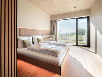 Helles Schlafzimmer mit großem Bett, Holzdekor und großem Fenster mit Blick ins Grüne.