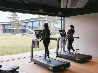 Zwei Frauen laufen auf Laufbändern in einem modernen, gut beleuchteten Fitnessraum mit Blick auf einen grünen Innenhof.