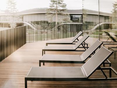 Drei graue Liegestühle auf einer Holzterrasse mit Glasgeländer und Bäumen im Hintergrund.