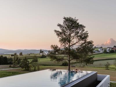 Moderner Infinity-Pool mit Blick auf eine grüne Hügellandschaft und einen Baum bei Sonnenuntergang.