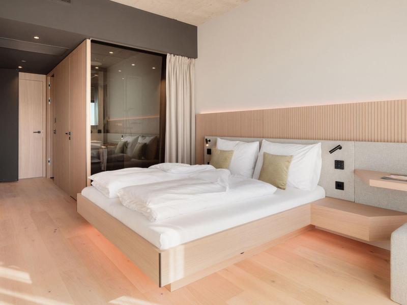 Modernes Schlafzimmer mit Doppelbett, Holzboden, hellem Design und Glaswand zur Dusche.