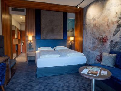 Modern eingerichtetes Hotelzimmer mit einem Doppelbett, Sofa und großflächiger Wanddekoration.