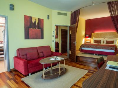 Modern eingerichtetes Hotelzimmer mit rotem Sofa, rundem Tisch und großzügigem Schlafzimmerbereich.