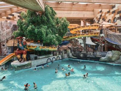 Belebtes Hallenbad mit Rutschen, Wasserattraktionen und Badegästen unter Holzdecken und Wanddekorationen.