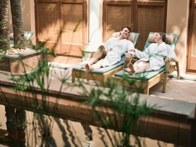 Zwei Personen relaxen auf Liegen in Bademänteln neben einem Pool vor einem Holzgebäude.