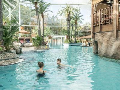 Großer Indoor-Pool mit Palmen und zwei Personen, umgeben von einer tropischen Hotelumgebung.