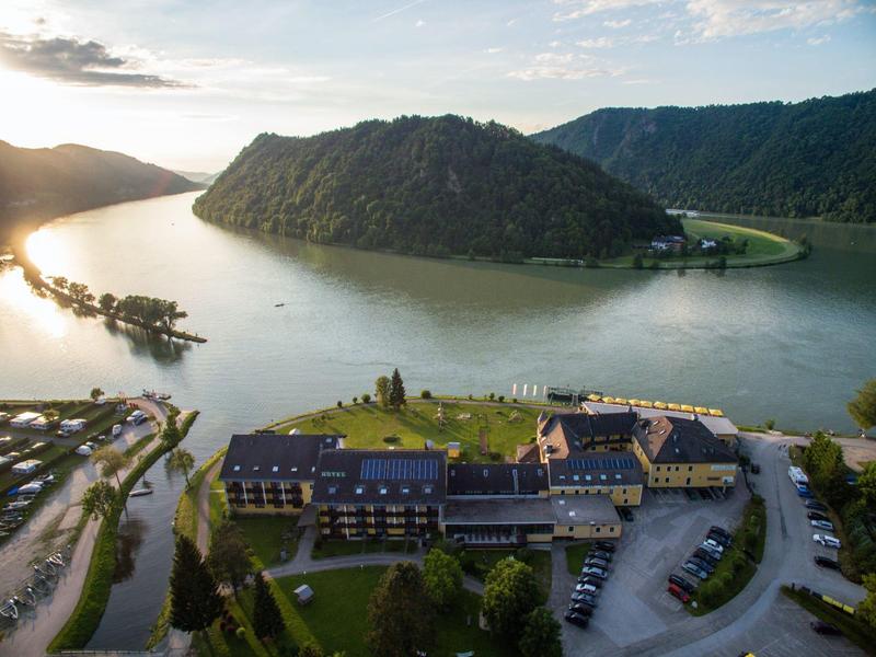 Fluss mit großer Halbinsel, umgeben von grünen Hügeln und mehreren Gebäuden am Ufer bei Sonnenuntergang.