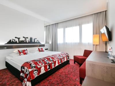 Modernes Hotelzimmer mit Doppelbett, rotem Teppich, Sessel, großem Fenster und Wand-TV.