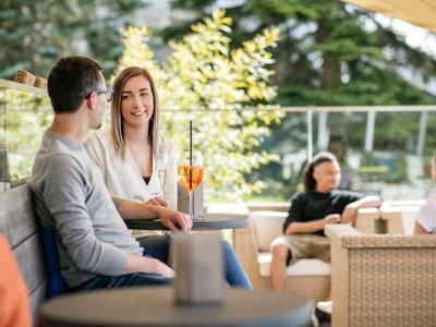Zwei Personen sitzen auf einer Terrasse und unterhalten sich bei Getränken, im Hintergrund weitere Gäste.