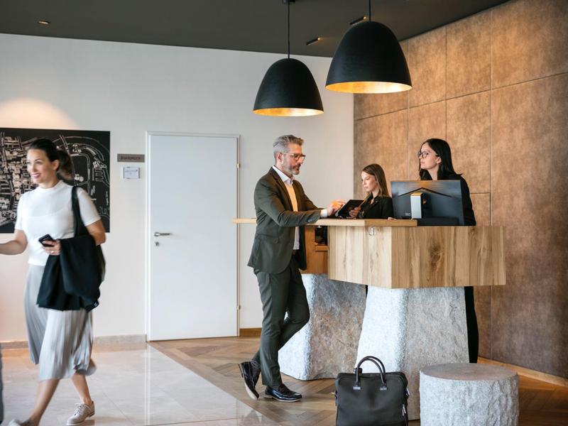 Ein Mann checkt am Empfang ein, zwei Damen am Schalter helfen ihm, eine Frau mit Koffer geht vorbei.