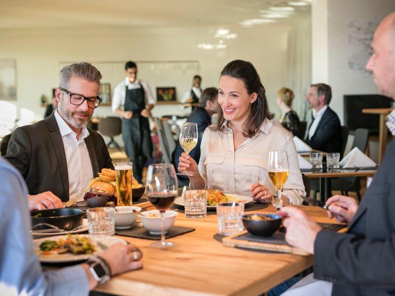 Vier Erwachsene sitzen an einem Holztisch, essen und trinken in einem hellen Restaurant.