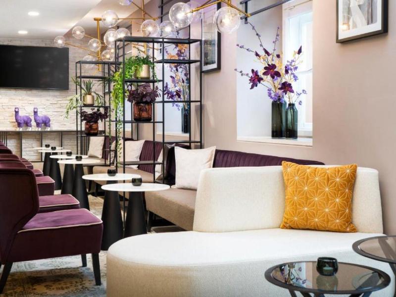 Area lounge moderna dell'hotel con sedie bianche e viola, scaffali in vetro e metallo, cuscini decorativi.