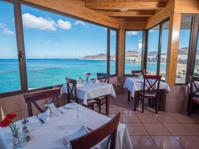 Restaurant mit Holzdach und weißen Tischtüchern, Blick auf blaues Meer und Himmel bei Sonnenschein.
