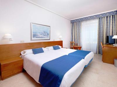 Camera d'hotel luminosa con letto matrimoniale, biancheria blu e bianca e finestra con tende.