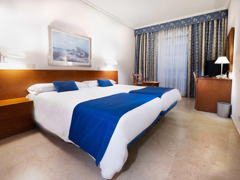 Camera d'hotel luminosa arredata con letto matrimoniale e coperta blu accanto a una finestra con tende.