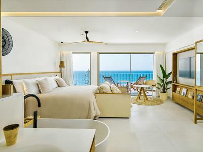 Helles, modernes Schlafzimmer mit Meerblick, Holzmöbeln und gemütlicher Sitzgruppe vor großen Fenstern.