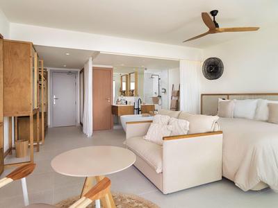 Helles Schlafzimmer mit weißem Bett, Sofa, rundem Tisch, Holzmöbeln und Deckenventilator.