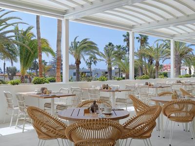 Freiluftrestaurant mit weißen Tischen, geflochtenen Stühlen und Palmen im Hintergrund.