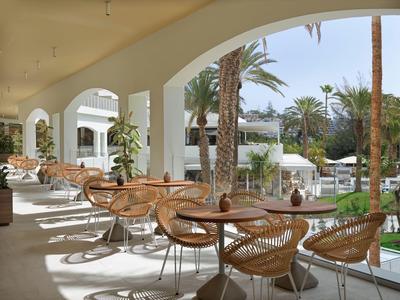 Überdachte Terrasse mit runden Tischen, geflochtenen Stühlen und Palmen im Hintergrund bei Tageslicht.