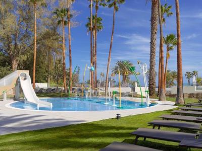 Piscina dell'hotel con scivoli d'acqua, lettini e alti palme sotto un cielo azzurro.