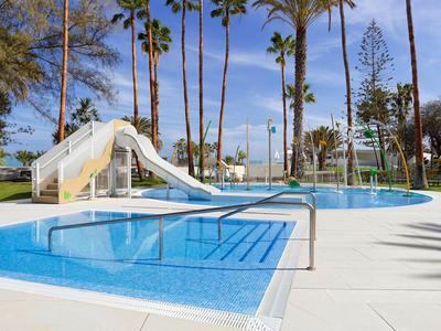 Piscina rotonda dell'hotel con scivolo, circondata da palme e sentieri chiari.